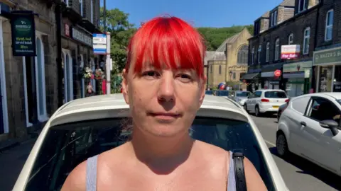 A woman with dyed red hair and grey strapped top standing in a main street with cars in the background.