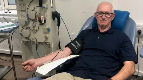 A man with glasses, a dark shirt and beige shorts, sits on a chair in a doctor's office while donating blood. The machine is standing next to him.