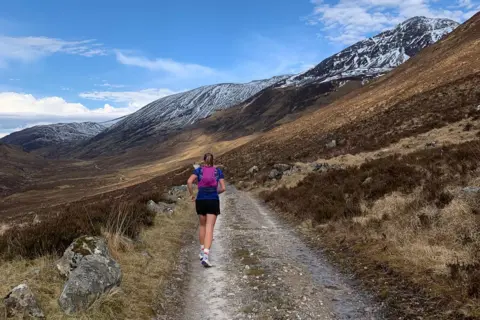 Freya Shepherd Freya in the middle of the shot running on the trail. She is running towards the mountains with their tops covered in snow. She is wearing a navy T-shirt, black shorts and magenta backpack
