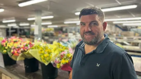 A man with dark hair and a short beard, wearing a navy polo, smiles at the camera. Behind him is a flower packing warehouse, with five bouquets lined up of yellow and pink flowers.