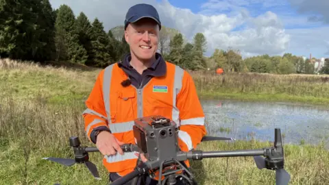 Duncan Turner smiles at the camera in a orange jacket and a blue cap. He is holding the drone. Behind him is a large pond in a field.