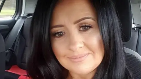 A woman is seen with long black hair and is smiling while sitting in the front seat of a car with black upholstery.She is wearing a red top.  