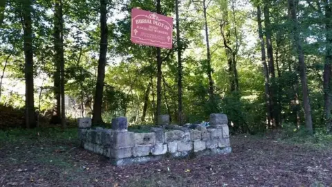 Duffy's Cut Project The picture shows a stone memorial in a forest. A red sign on the memorial reads: "Burial plot of Irish railroad workers".