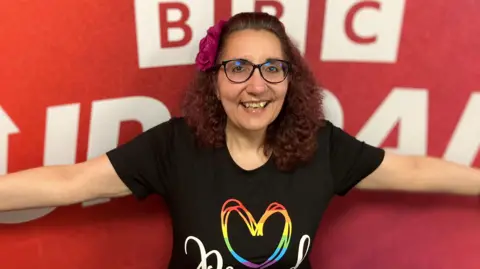 A woman in a black t-shirt with a multi-coloured heart design on the front stands in front of a red BBC backdrop with her arms stretched wide. She has curly shoulder-length plum-coloured hair and wears a burgundy flower to the side of her temple. She is wearing brown-framed glasses and smiling widely. 