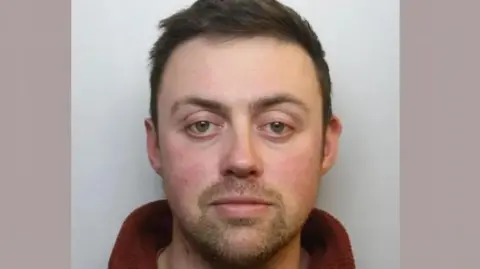 Wiltshire Police Mug shot of Andrew Edwards. He is in his early to mid thirties with a trimmed beard and short brown hair, he has slightly red eyes and cheeks and wears a brown fleece with the zip open.