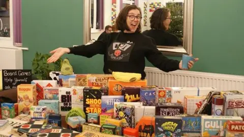 @cardsordie/Instagram Ann, with short black hair and glasses and a black "cards or die" jumper on. She is smiling and gesturing her arms to the table packed with about 60 games in front of her.