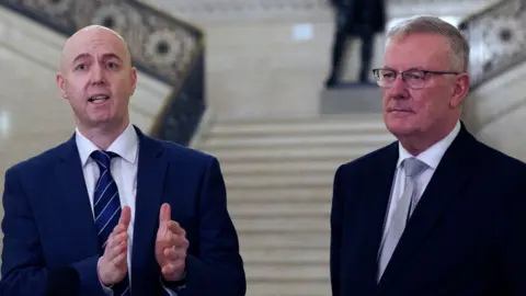 Jon Burrown and Mike Nesbitt. Jon os bald, wearing a white shirt and blue suit. Mike has short grey hair, wearing a white shirt, grey tie and black blazer.
