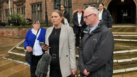 Claire Chetwynd, Mr Carpenter's stepdaughter, speaking after his inquest. She has a smart coat on and is holding a phone with a microphone in front of her. She is surrounded by family. 