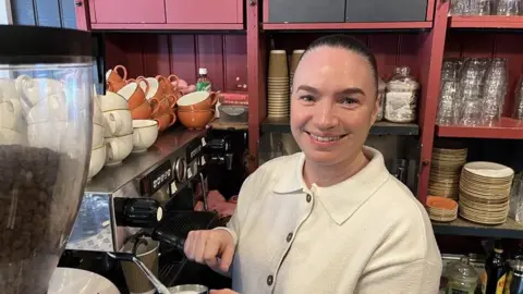 Phil Harrison / BBC A woman wearing a white top stood in a pink cafe next to a silver barista-style coffee machine.