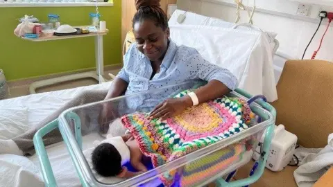 A mum sits on a hospital bed looking into a plastic cot that contains a baby wrapped in a colourful knitted blanket.