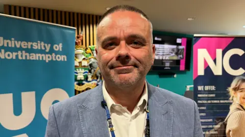 Ollie Conopo/BBC A man in a light blue suit with a white open-necked shirt stands in front of a blue banner. He is smiling at the camera.
