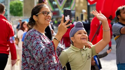 Bradford Council A woman and young boy smiling as they look up at something and take photos. They look like a mother and son. She wears a patterned red and blue top, and he wears a tan T-shirt and a child-size turban. They are surrounded by people milling around in the background.