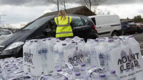 An image of packs of bottled water stacked up. Behind the bottles is a person with long blonde hair in a high-vis vest talking to someone through the window of a black car