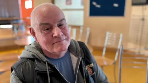 A man sits in a leisure centre. He has grey hair and is smiling at the camera. His brows and raised. He's dressed in a t-shirt, with a zip up hoodie and a body warmer on top. behind him are some chairs and a noticeboard.