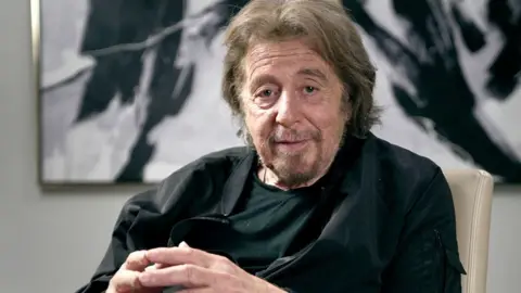 BBC Al Pacino sits and looks directly at the camera, his fingertips touching, with a piece of art behind him on the wall.