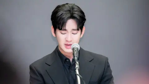 iMBC / Imazins via Getty Images A tearful Kim Soo-hyun looking down and speaking into a microphone. He is wearing a black shirt and black blazer. There is bokeh in the foreground.