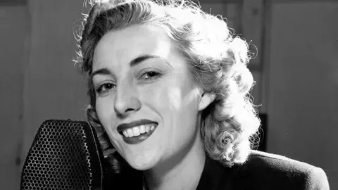 A black and white photograph shows Vera Lynn on her war time radio programme, Sincerely Yours. She has wavy hair and is wearing makeup and lipstick and is sitting in front of a microphone smiling at the camera.
