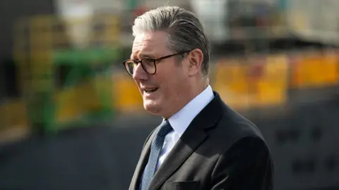 Prime Minister Sir Keir Starmer speaks at an outdoor press conference wearing a black suit, white shirt and navy blue tie.