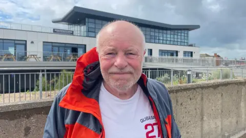 David Andrews is a man in his 70s, he is bald with a grey stubble beard. He is standing in front of the Seaview Colonnade building. He is wearing a grey and orange waterproof jacket, with a white T-shirt underneath. 