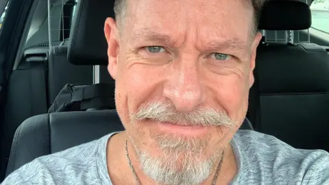 Antony Loveless Antony Loveless sits in his car. You can see his head and his shoulders and he has a grey beard and moustache. He is wearing a silver chain and has a grey t-shirt. The car has black seats. He is grinning at the camera and has green/grey eyes.