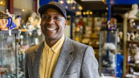 Wearing a baseball cap and a yellow shirt with a grey blazer over the top Ochuko Ojiri looks directly at the camera smiling. In the background are dozens of antiques displayed around him