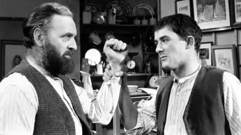 Andrew Keir, a bearded man with an intense look about him, is wearing a grandad shirt and tweed waistcoat. He has lifted his hand aggressively and it has been grabbed by the actor Paul Young, equally intense but not bearded, wearing a similar shirt and waistcoat. Behind them is a welsh dresser filled with kitchenware and a wall covered in pictures.