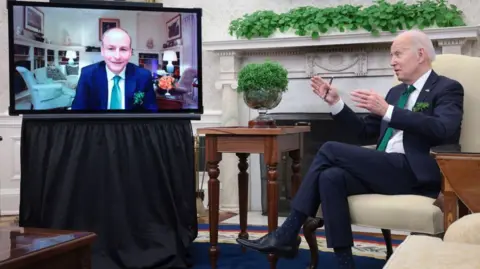 Getty Images Micheál Martin is seen on a TV screen in the Oval Office in a blue suit and green tie, next to the TV is a bowl of shamrock on a mahogany table and next to that is Joe Biden in a dark suit, white shirt and green tie.