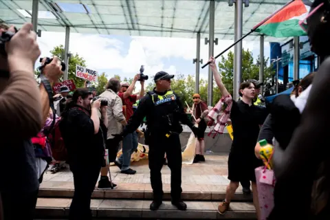 Cason Ho / ABC News A police officer looks on as protesters wave flags and signs - one saying 'no pride genocide' and a Palestinian flag in view - in Perth on Monday.
