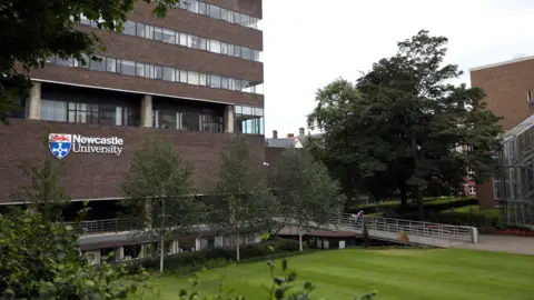 NCJ Media A Newcastle University campus. A dark brick brutalist building with narrow windows sits to the left. There is a sign on it which reads Newcastle University alongside the institution's crest. In front of the building there is grass and trees.