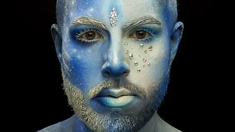 Rahual Das Rahual has a full-face make-up look and is looking into the camera. His entire face and shoulders have been painted in different hues of blue. There are white highlights and accents, such as paint splatters on one side of the face that look like a cluster of stars. His eyelashes, eyebrows and beard are painted white. In between his brows is a sparkly bindi.