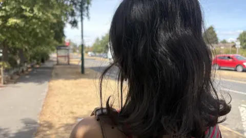 The back of an anonymous woman who has shoulder-length black hair. She is wearing a grey and red off-the-shoulder top