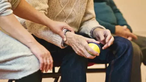 Partial view of three seated people, showing hands, arms and knees. The man in the middle appears to be elderly and is wearing jeans, a cable knit jumper and holding a small coloured ball in one hand. To his right a younger woman has her hand on his wrist. She is wearing a long grey jumper with the sleeves pushed up. The person in the left, and slightly out of focus, is wearing a dark green jumper and has their hands clasped on their lap.