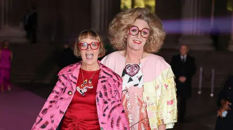 Getty Images Philippa Perry and Grayson Perry on a red carpet and both wearing brightly coloured clothes. Philippa is wearing a red dress with a pink and black patterned coat and is smiling at the camera with her mouth open.