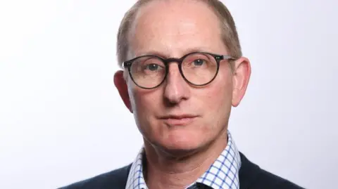 Ormiston Families Bruce Leeke looks directly at the camera with a neutral expression. He is wearing a blue and white checked shirt and dark-coloured jacket and dark-framed glasses.
