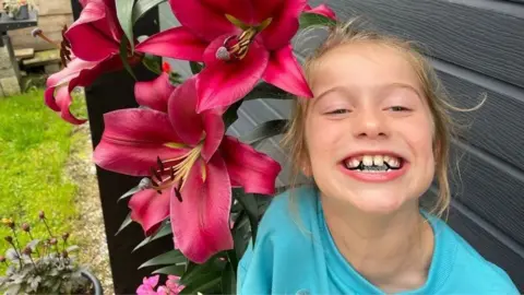 Family Handout Scarlett Rossborough smiling beside large pink lily