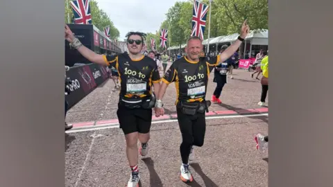 Joel Pennington Beau and Gary McKee hold hands as they cross the finish line at the London Marathon. They are both wearing black and yellow tops that read: "100 to LDN". Their free hands are raised in the air. There are union jack flags erected down the Mall behind them.