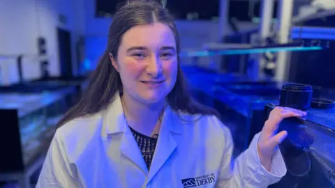Triona Barker in a white lab coat stands beside her lab experiments