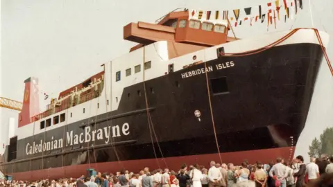 This is a picture of crowds gather at Cochrane's Shipyard in Selby to watch the 1985 launch of a newly built ship, Hebridean Isles. The main part of the ship is painted blue - the name of its owners - Caledonian MacBrayne - is painted on the side in white. Flags of various nations are hanging above the ship.