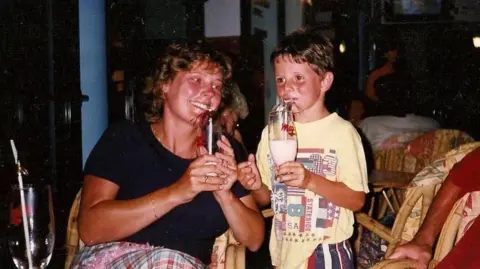 Diane Taylor and her son Daniel as a young child in a family holiday photo - they are smiling and sipping on drinks together.