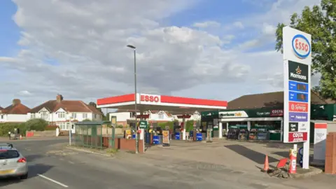 An Esso fuel station forecourt with a Morrisons Daily store. The petrol station is situated on the corner of two roads. There are no cars in the forecourt and houses can be seen on the other side of the road. It is a partly cloudy day with patches of blue sky.