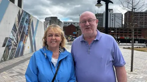 A photo of a man and woman outside Belfast Grand Central Station. The woman Rosie on the left is wearing a white top with a blue rain jacket; she has short blonde hair.On the right is a man Patrick, wearing a purple polo shirt and glasses.