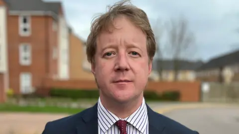 BBC Paul Bristow stares at the camera. He wears a dark suit, pinstripe shirt and red tie with white spots. He stands in a residential street. 