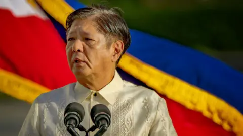 Getty Images Wearing a white linen shift, Ferdinand "Bongbong" Marcos Jr speaks into a microphone at a rally in front of the Philippine flag