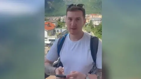 Submitted A man with short brown hair with sunglasses on his head has a serious look on his face. He is wearing a white top and backpack and has tattoos on his arm. He is holding a phone and in the background you can see several houses and a mountain in the distance.