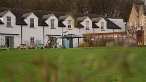 The outside of a hospice. It's a row of terraced white townhouses, with picnic tables outside and a large lawn.