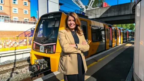 North East Combined Authority North East Mayor Kim McGuinness standing on a train platform with her arms crossed. She has light brown hair and is smiling. She is wearing a camel-coloured coat. A yellow-and-grey Metro train is behind her on the platform.