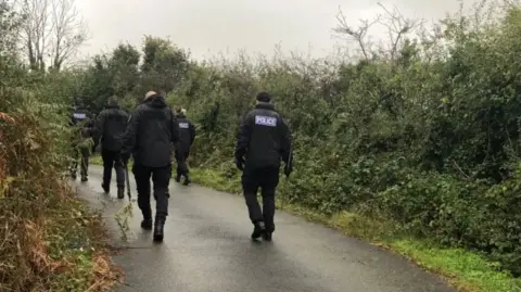 BBC Police officers wearing black coats, some with the 'police' logo on the back, walking along a Cornish lane bordered by hedgerows