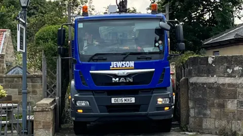 Nerys Mitchell A blue Jewson lorry backing down a small residential side-street. Nerys is driving and looking in her wing mirror, with one hand on the wheel. She is wearing a dark-coloured top and a yellow hi-vis vest.