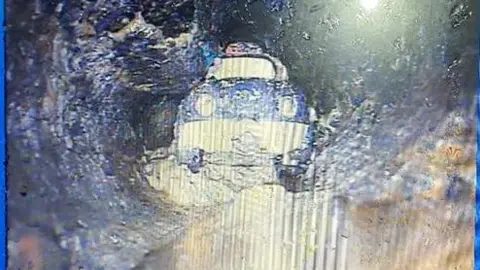 The toy car pictured in a drain. It appears to be a blue and white plastic car and is sitting in sludge.