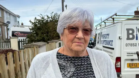 Lady with grey hair and glasses looks at the camera whilst stood in front of a maintenance van and to the right of a wooden fence